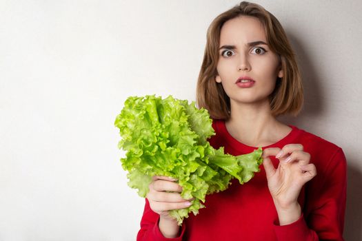 Concerned brunette woman wears red sweater holding lettuce over a grey background. Empty space