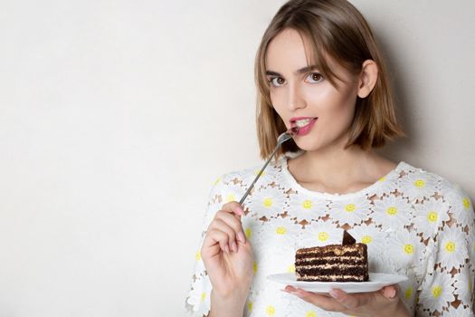 Thoughtful young woman wearing white dress with flowers eating tasty cake with cream. Empty space