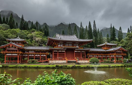 Kaneohe, Oahu, Hawaii, USA. - January 11, 2020: Maroon walls and gray roof, Byodo-In Buddhist temple with koi-pond in front, green trees and mountain range in back under dark rainy sky.