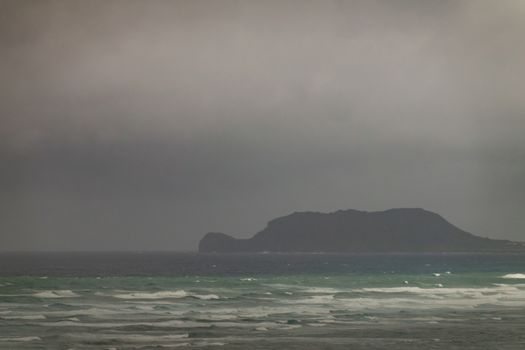 Kaaawa, Oahu, Hawaii, USA. - January 11, 2020: Mokolii Island in Pacific Ocean azure and white surf with gray water towards horizon under gray rainy clouds off Kaaawa on East Coast of island.