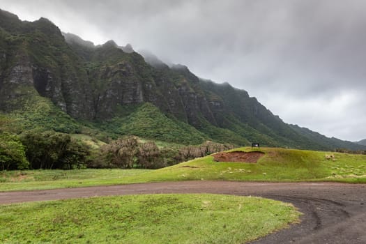 Kaaawa, Oahu, Hawaii, USA. - January 11, 2020: Dirt road makes U-turn ingreen meadow with dark brown to black high rocky cliffs on side of Kualoa valley under cloudy rainy sky.