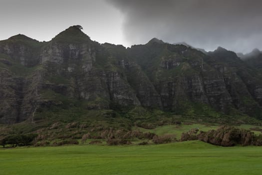 Kaaawa, Oahu, Hawaii, USA. - January 11, 2020: High dark brown to black rocky cliffs of mountain range border Kualoa valley under cloudy rainy sky.