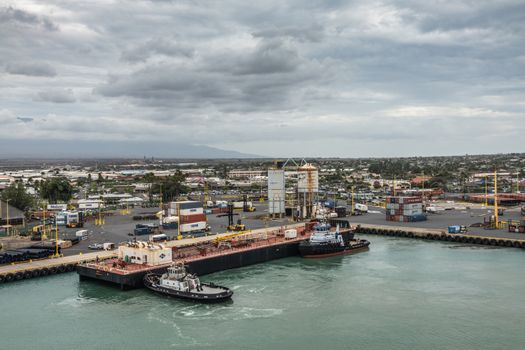 Kahului, Maui,, Hawaii, USA. - January 13, 2020: Nale double hull liquid tank barge pushed by 2 tugboats against container quay in port under rainy cloudscape. Buildings of town in back.