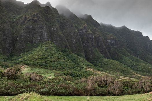 Kaaawa, Oahu, Hawaii, USA. - January 11, 2020: Brown to black high rocky cliffs of partly green forested mountains on side of Kualoa valley under cloudy rainy sky.