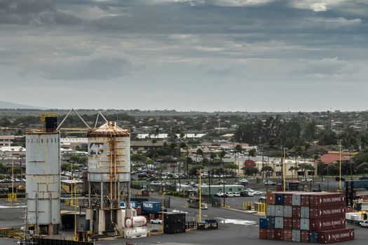 Kahului, Maui,, Hawaii, USA. - January 13, 2020: Tall metal fuel tanks and stacks of shipping containers in front of cars and green cityscape under thick gray-blue cloudscape.