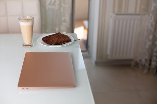 Closed laptop with a glass of coffee and homemade chocolate pie on the table. Study at home concept. Space for text