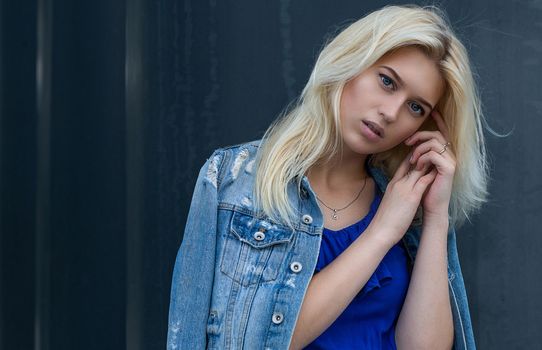 Street photosession of pretty blonde girl wears casual apparel posing near the grey wall. Space for text