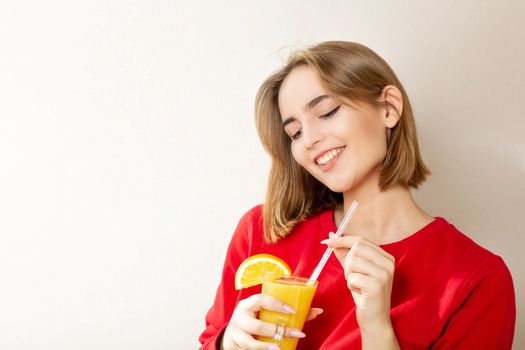 Pleased young woman wears red sweater holding glass of orange juice over a grey wall. Space for text