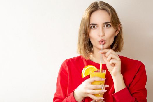 Surprised young woman wears red sweater drinks orange juice over a grey wall. Space for text