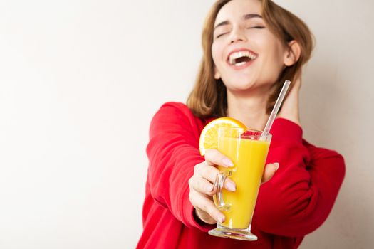 Merry young woman wears red sweater holding glass of orange juice over a grey wall. Space for text