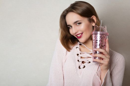 Cheerful young woman wears pink sweater holding glass of orange juice over a grey wall. Space for text