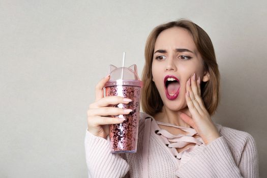 Surprised young woman wears pink sweater holding glass of orange juice over a grey wall. Space for text