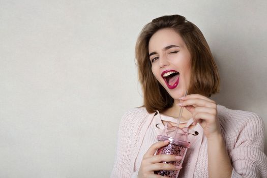 Winking young woman wears pink sweater holding glass of orange juice over a grey wall. Space for text