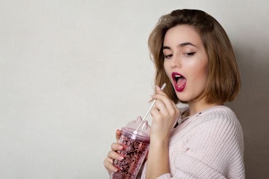 Emotional young woman wears pink sweater holding glass of orange juice over a grey wall. Space for text