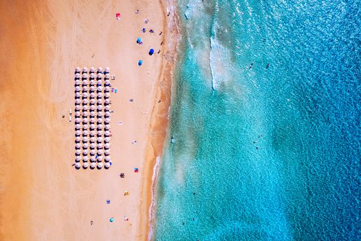 Aerial view of sandy beach with colorful umbrellas, swimming people in sea bay with transparent blue water at sunset in summer. Aerial top view on the beach, umbrellas, sand and sea waves.
