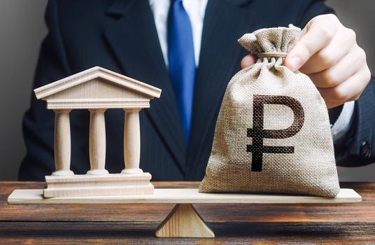 A man puts a russian ruble money bag on scales opposite to government building, bank, university. State debt. Budget balance. Taxes payment. Lobbying interests. Loans. Reforms financial support.