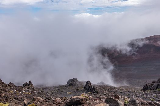 Haleakala Volcano, Maui, Hawaii, USA. - January 13, 2020: Whtie fog lingers in crater. Brown, and black rocks and yellowish ground vegetation at edge of crater. Blue patches in sky.