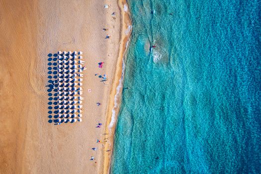 Aerial view of sandy beach with colorful umbrellas, swimming people in sea bay with transparent blue water at sunset in summer. Aerial top view on the beach, umbrellas, sand and sea waves.