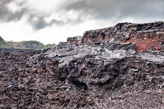 Leilani Estate, Hawaii, USA. - January 14, 2020: 2018 Kilauea volcano eruption hardened black lava field. Thick layer with cliffs and brown and red colors under rainy sky and green forest on horizon.