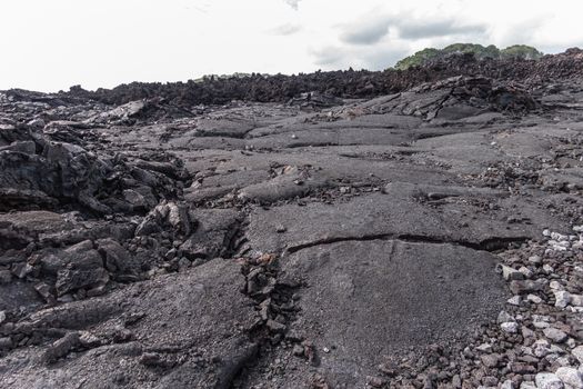 Leilani Estate, Hawaii, USA. - January 14, 2020: Large 2018 Kilauea volcano eruption hardened black lava field under white cloudscape and some green trees on horizon peeping above.