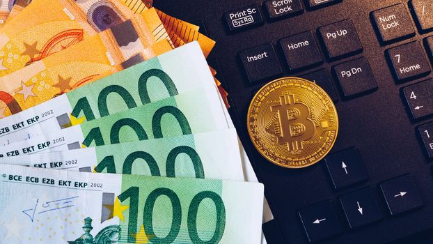 Golden bitcoin over Euro money. Bitcoin cryptocurrency. Crypto currency concept. Bitcoin with euro bills. Bitcoins stacked on euro banknotes. Computer keyboard used as background.