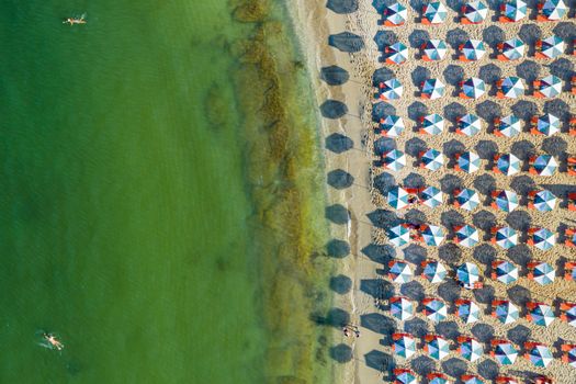 Aerial view of sandy beach with colorful umbrellas, swimming people in sea bay with transparent blue water at sunset in summer. Aerial top view on the beach, umbrellas, sand and sea waves.