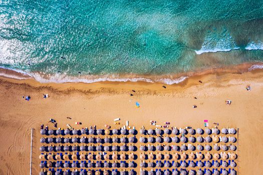 Aerial view of sandy beach with colorful umbrellas, swimming people in sea bay with transparent blue water at sunset in summer. Aerial top view on the beach, umbrellas, sand and sea waves.