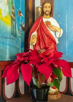 Kalapana, Hawaii, USA. - January 14, 2020: Mary, Star of the Sea Catholic Church. Closeup of Sacred Heart Jesus statue with white and red dress behind red flowers. Blue walls and brown beams in back.