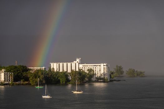 Hilo, Hawaii, USA. - January 14, 2020: Rainbow in dark rain and storm sky touches Grand Naniloa Hotel on shore. Sun on white walls and sides of 3 sailing boats. Green foliage.