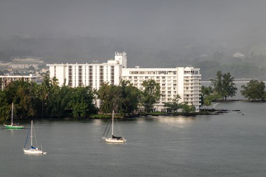 Hilo, Hawaii, USA. - January 14, 2020: Dark rain and storm sky above closeup of Grand Naniloa Hotel on shore. Sun on white walls and sides of 3 sailing boats. Green foliage.