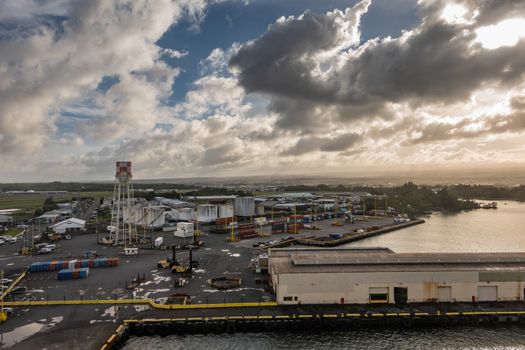 Hilo, Hawaii, USA. - January 14, 2020: Wide shot over most of local port with tanks, stacked shipping containers, quays, warehouse, trucks, cars and more. All under brown cloudscape with blue patches.