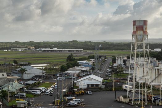 Hilo, Hawaii, USA. - January 14, 2020: Road into ocean port with warehouses and tanks on both side. Tall water tower. Green airport under gray cloudscape. Green belt on horizon.