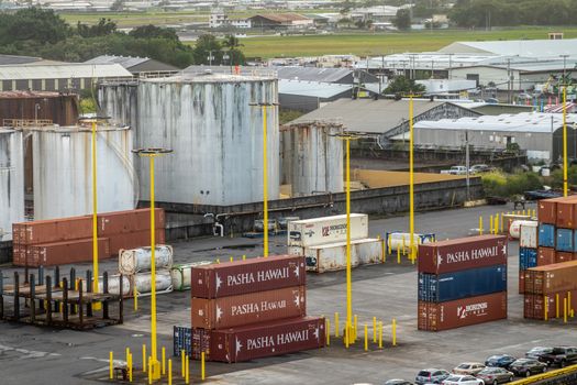 Hilo, Hawaii, USA. - January 14, 2020: Closeup of stacks of maroon Pasha shipping containers in front of gray fuel tanks. Green of airport with control tower in back.