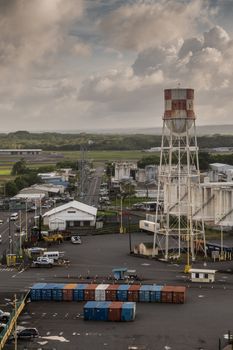 Hilo, Hawaii, USA. - January 14, 2020: Access road to ocean port with water tower and line of shipping containers under heavy brown cloudscape