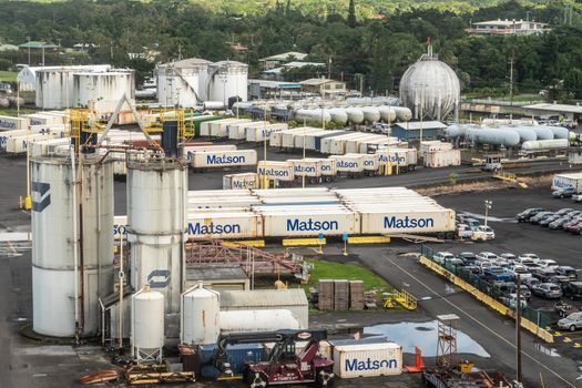 Hilo, Hawaii, USA. - January 14, 2020: Ocean port. White gas and fuel tanks surrounded by Matson shipping containers placed on trailers. Green tree on horizon. Cars and other stuff.
