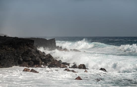 Kaimu Beach, Hawaii, USA. - January 14, 2020: Azure wave crashes with white surf on black lava coastline under heavy rain brown cloudscape. 