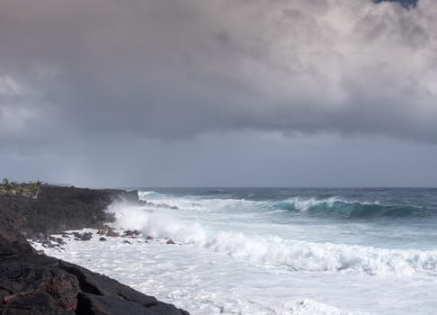 Kaimu Beach, Hawaii, USA. - January 14, 2020: Azure wave crashes with white surf on black lava coastline under heavy rain brown cloudscape. 