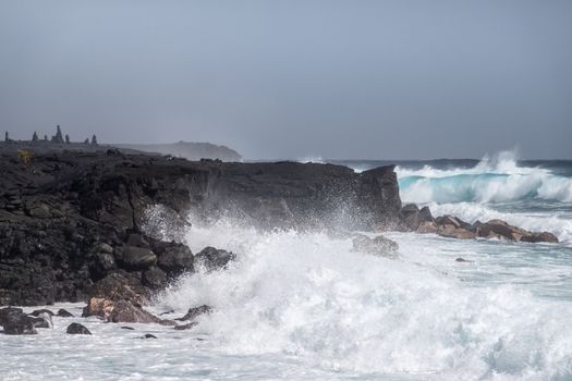 Kaimu Beach, Hawaii, USA. - January 14, 2020: Violent Azure waves crash with white surf on black lava coastline under heavy rain gray cloudscape. 