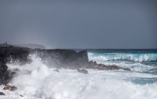 Kaimu Beach, Hawaii, USA. - January 14, 2020: Violent Azure waves crash with white surf on black lava coastline under heavy rain gray cloudscape. 