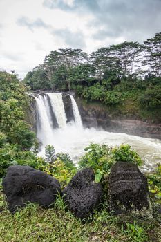 Hilo, Hawaii, USA. - January 14, 2020: White Rainbow Falls on foaming violent Wailuku River surrounded by green trees and plants under white-gray cloudscape.