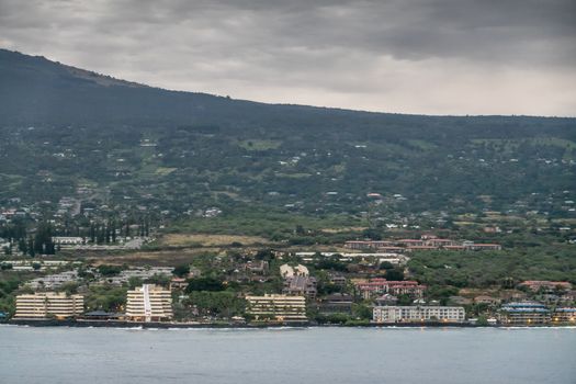 Kona, Hawaii, USA. - January 15, 2020: Resort hotel buildings along coastline set in green trees with forested mountain in back, dotted with red or white house roofs. Gray ocean in front.