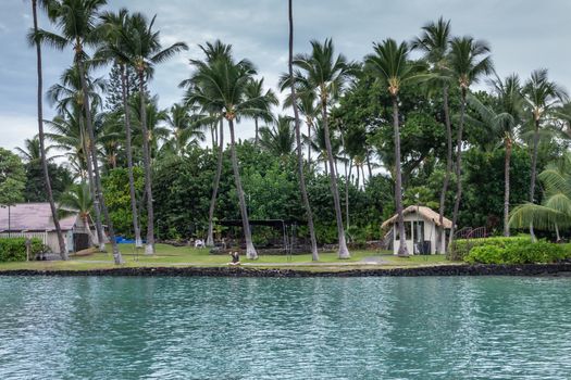 Kona, Hawaii, USA. - January 15, 2020: Kamakahonu Historic park. Meditating and praying on shoreline under palm trees and blueish cloudscape. Lots of green palm trees and small hut-like constructions.