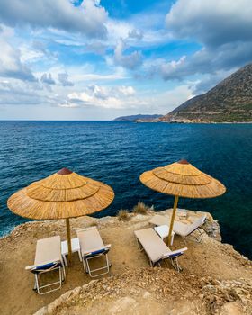 Summer vacation destination. Straw sunshades and sunbeds on the empty rocky beach with sea in the background. Vacation And Tourism Concept. Sunbeds On The Paradise Beach. Umbrellas and sunbeds.