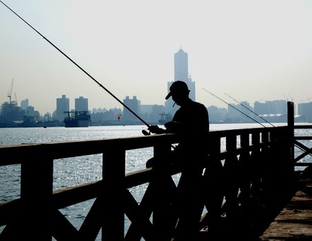KAOHSIUNG, TAIWAN -- SEPTEMBER 12 , 2017: A fisherman checks his tackle near the port entrance. In the back is the skyline of Kaohsiung City.
