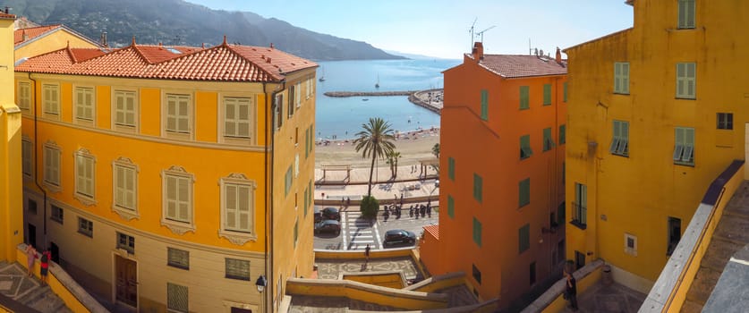 Menton, France - June 30, 2018: Bright facades of colorful houses and the blue sea on a Sunny day. Menton is a small seaside town on the French Riviera. Some people standing on a viewpoint.