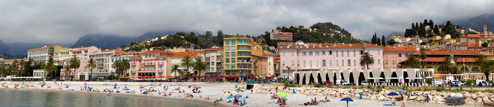 Menton, France - June 30, 2018: Menton beach and a panoramic view of the city. Unidentified people rest on the shore. Menton is a small seaside town on the French Riviera.