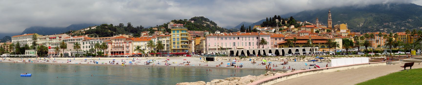 Menton, France - June 30, 2018: Menton beach and a panoramic view of the city. Unidentified people rest on the shore. Menton is a small seaside town on the French Riviera.