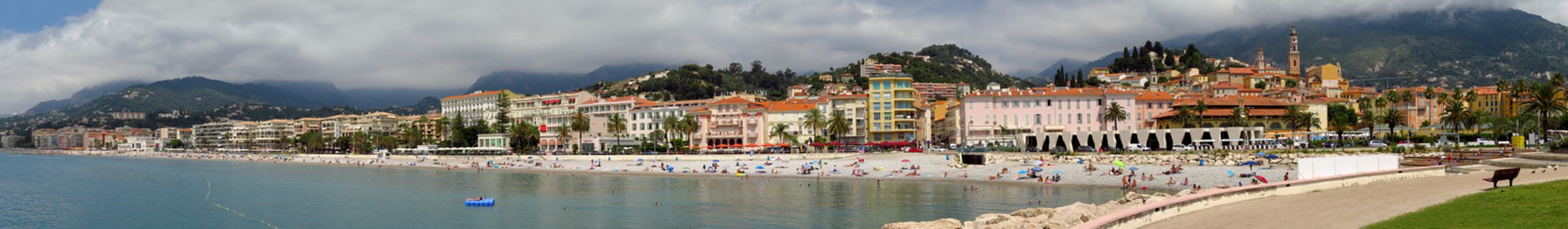 Menton, France - June 30, 2018: Menton beach and a panoramic view of the city. Unidentified people rest on the shore. Menton is a small seaside town on the French Riviera.