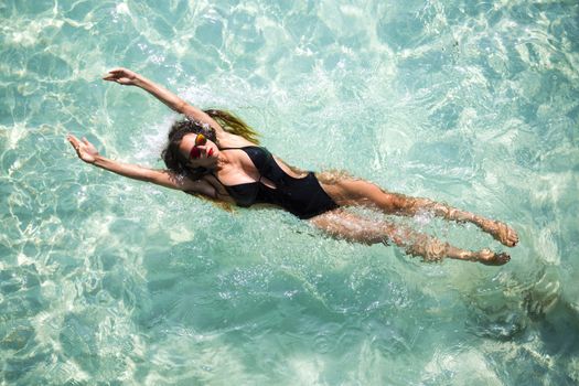 Young woman wearing a black monokini is diving in the clear water on her back in the Maldives. This creative photo has been taken from the top of a bungalow. Mirror sunglasses and red lipstick is being weared by the lady.