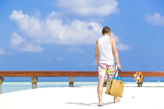 Man walking by the shore in the beach going away to a jetty. He is wearing a colorful bathing suit while carrying a beach bag with towels in  it. At background there is a beautiful sea with different tones of turquoise out of focus.